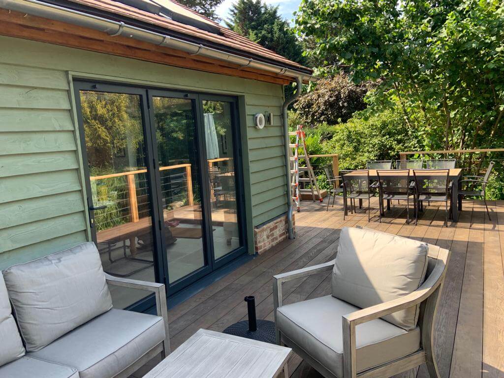 Outdoor deck area featuring comfortable seating and a dining table, surrounded by greenery and large glass doors.