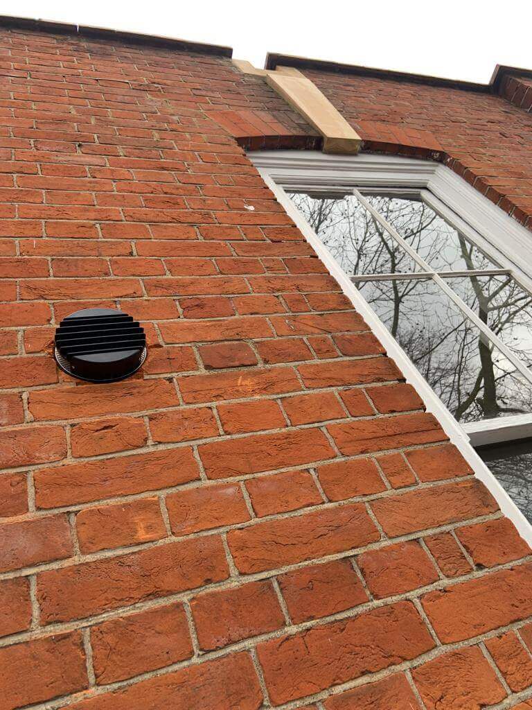 Exterior view of a brick wall with a black ventilation grille, showcasing a heat recovery ventilation system installation.