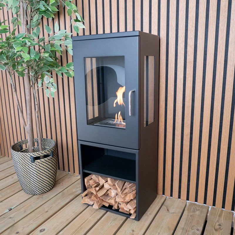 Cheyenne freestanding bioethanol fireplace with glass sides and wood storage, set against a wooden wall.