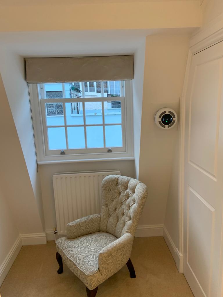 Cozy corner with a patterned armchair, window with a blind, and a heat recovery ventilator on the wall.