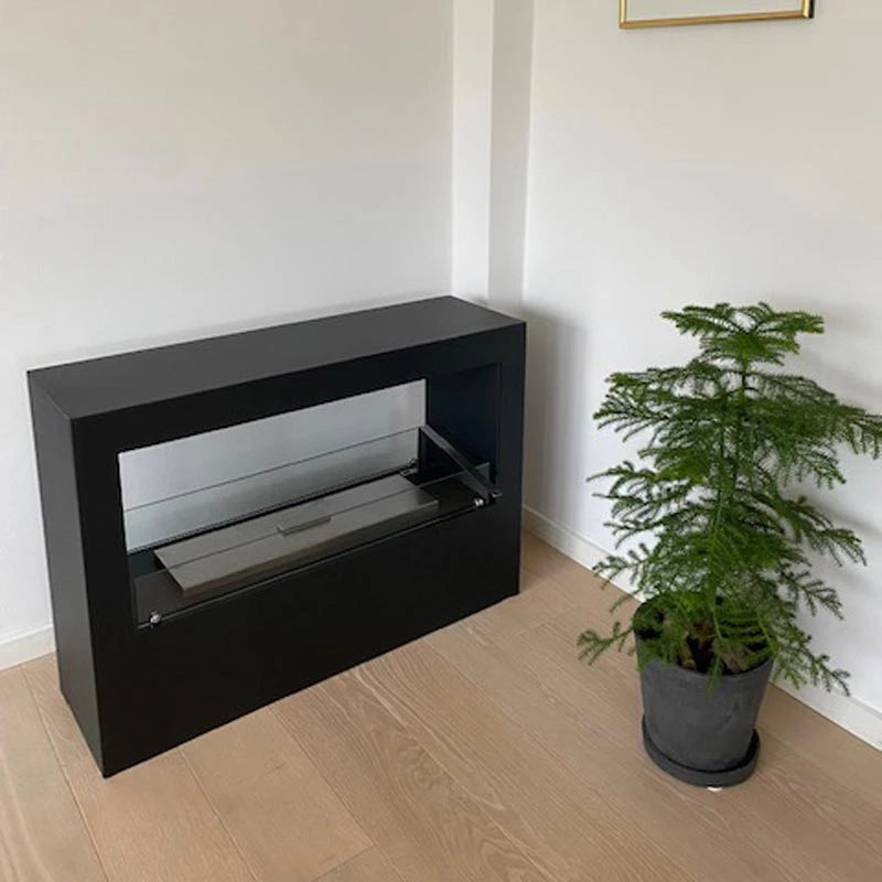 Lansing Tunnel Bioethanol Fireplace in black, featuring a glass front, serving as a stylish room divider with a plant beside.