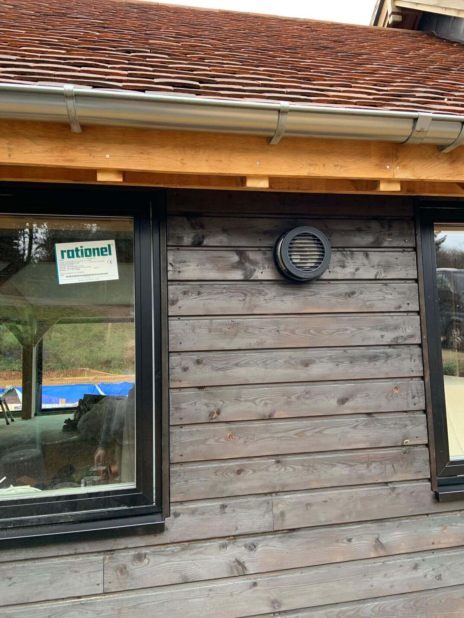 Exterior view of a wooden building featuring a round ventilation grill, showcasing a heat recovery ventilation system.