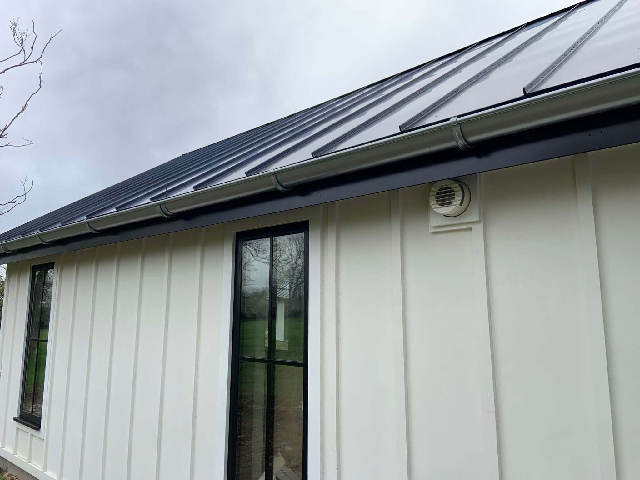 Side view of a modern house showcasing a metal roof, large windows, and a heat recovery ventilation outlet.