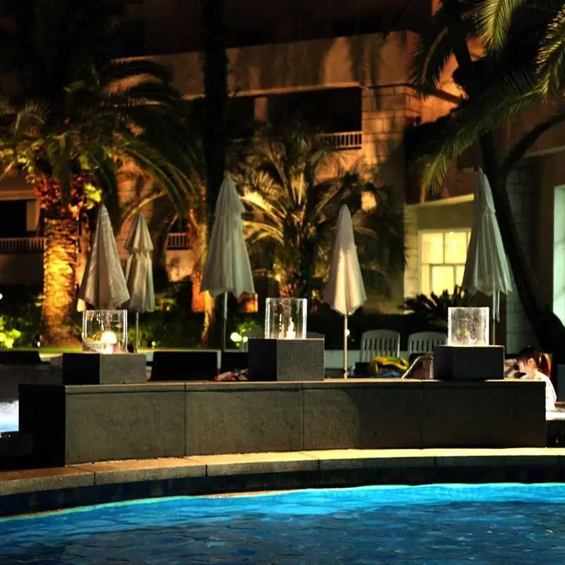 Modern bio-ethanol fireplace inserts by the poolside, surrounded by palm trees and outdoor umbrellas, at night.
