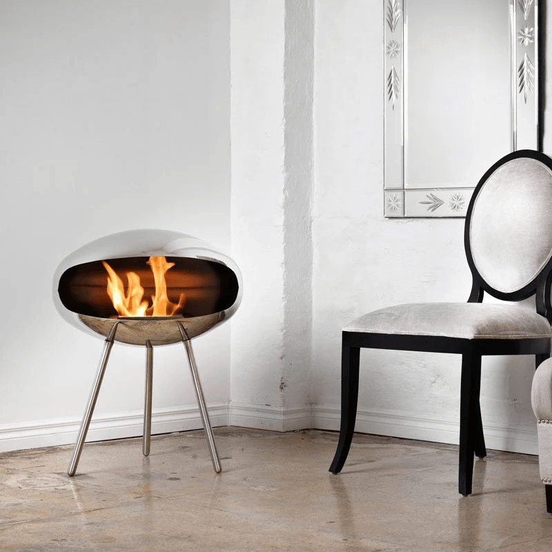 Cocoon Terra modern bioethanol fireplace in polished steel, featuring a stylish design and flames, placed beside a chair.
