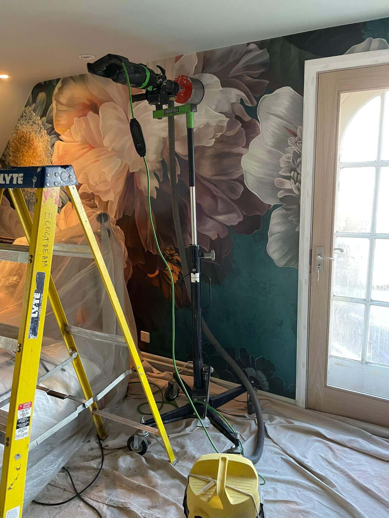 Installation process in a room featuring a floral wall mural, ladder, and equipment for ventilation or painting.