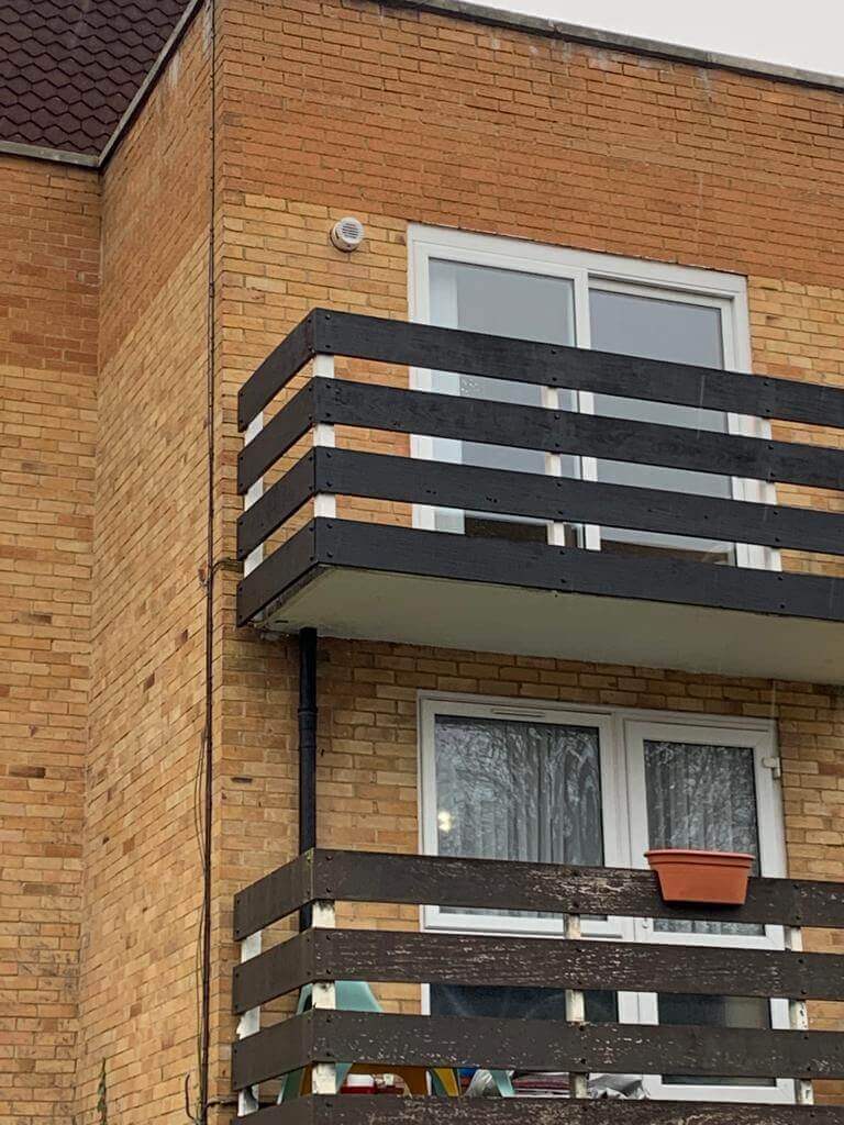Exterior view of a brick building with a balcony, featuring black wooden railings and a potted plant.