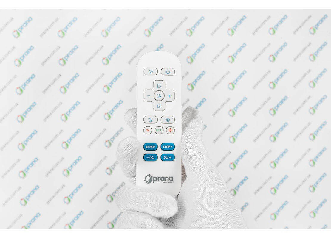 Hand holding a Prana remote control for heat recovery ventilation system on a branded background.