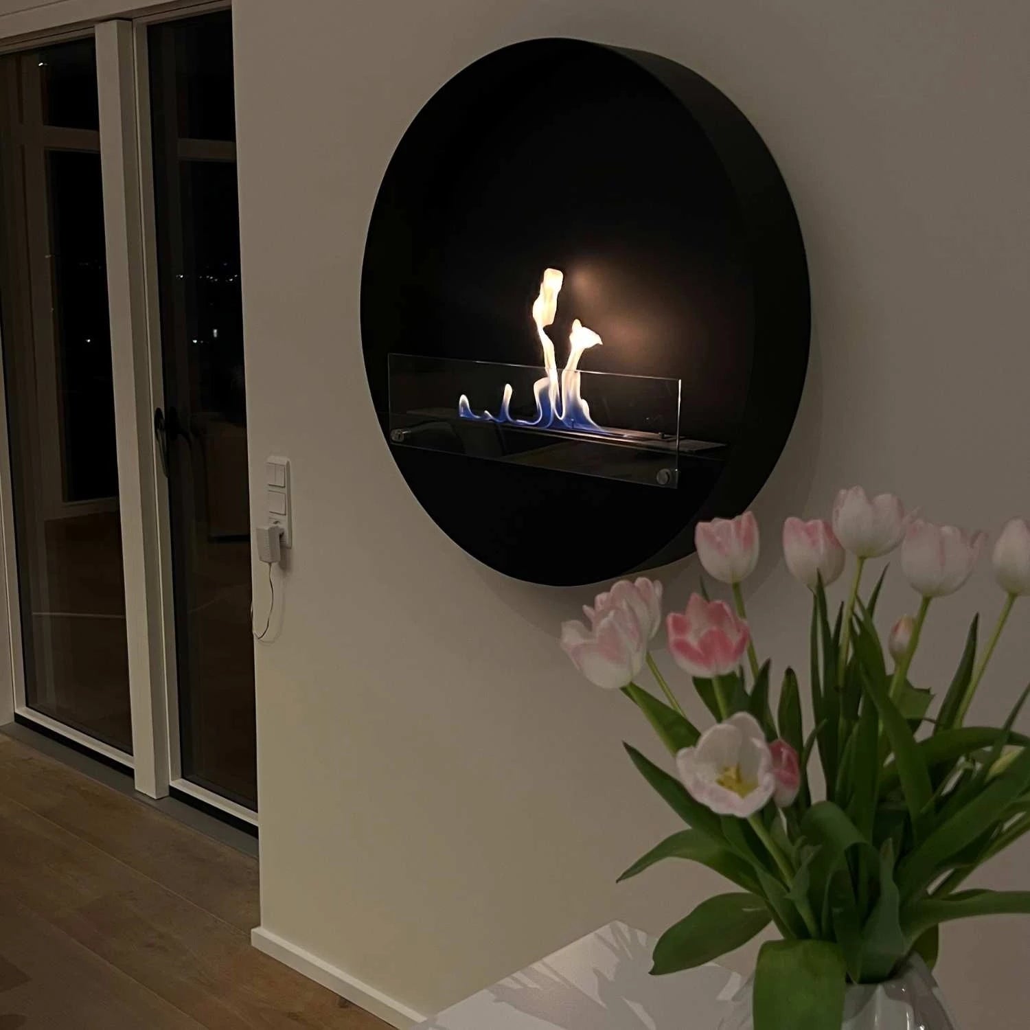 Delaware XL black round fireplace on wall, featuring flames and modern design, with flowers in foreground.