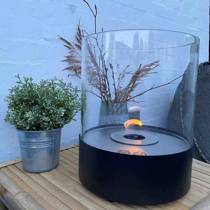 Southwark round tabletop bioethanol fireplace in black with clear glass, surrounded by greenery on a wooden surface.